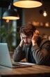 © Raisa - Man looks stressed at laptop screen, holding head in hands. He is in business casual attire, sitting at a wooden table with a document. Background shows warm lighting and decor.