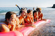 © Xavier Lorenzo - Diverse young friends are joyfully laughing and relaxing on a pink inflatable raft in the ocean water, experiencing a fun summer vacation at the beach during sunset