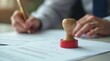 © Alex Pios - Hand with pen signs legal paper near red rubber stamp. Official document signing process, business agreement or contract. Businessperson completes paperwork with notary approval.