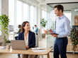 © Meow Creations - Woman working on laptop at office desk with male colleague standing nearby