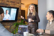 © Ina - Business meeting with four person. Woman presenting ideas while colleague joins via video call on screen. Concept of remote work, hybrid communication, collaboration and modern business environment.