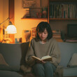 © AS_SleepingPanda - A serene young woman enjoys a quiet evening indoors, relaxing on a comfortable sofa and deeply focused on reading her book under the soft, warm lamplight in a cozy home environment
