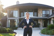 © Anastasiia - A businesswoman stands confidently in front of a luxurious, modern home. She is posing for a professional portrait. The setting is a well-maintained residential property with lush landscaping and sunn