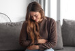 © luciano - Young and beautiful sick woman sits at home clutching her aching stomach, suffering from abdominal discomfort caused by indigestion, gastritis, menstrual cramps, or digestive problems, cramps