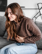 © luciano - Young and beautiful sick woman sits at home clutching her aching stomach, suffering from abdominal discomfort caused by indigestion, gastritis, menstrual cramps, or digestive problems, cramps