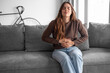 © luciano - Young and beautiful sick woman sits at home clutching her aching stomach, suffering from abdominal discomfort caused by indigestion, gastritis, menstrual cramps, or digestive problems, cramps