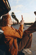 © SHOTPRIME STUDIO - Young woman with curly hair wearing orange jacket and brown pants smiles while sitting outdoors near car using smartphone in casual lifestyle setting during sunset.