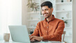 © Prostock-studio - A young man smiles while typing on a laptop at a table. He is in a bright room with plants in the background. A coffee cup is on the table beside him.