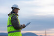 © 24K-Production - A young male electrical engineer works outdoors, inspecting high-voltage power lines with a tablet in a utility area at dusk.