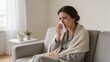 © olga_demina - A woman sits on a couch blowing her nose into a tissue while wrapped in a blanket, conveying a sense of illness and discomfort in a cozy indoor setting.