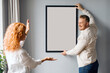 © Home-stock - European spouses hanging poster in frame on wall decorating living room together after renovation. Interior design concept