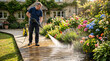 © Ahsan - A focused man stands firmly, efficiently pressure washing a wide wooden deck surface in a picturesque, well-maintained garden flourishing with beautiful flowers and plants.