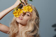 © VICHIZH - Beautiful young woman with long blonde hair holding yellow flowers, conveying a serene and artistic mood against a soft blue background.