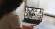 © fizkes - Businesswoman engages in virtual meeting with diverse group displayed on laptop screen, view over her shoulder. Integration of technology for professional communication in globally connected workspace