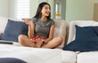 © wavebreak3 - Indian woman sitting cross-legged on sofa in living room wearing gray T-shirt using tablet