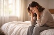 © justjupiter - A woman sits on the edge of a bed, holding her head in distress, in a softly lit bedroom.
