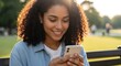 © Lerivan - Young woman with curly hair smiling while looking at her smartphone outdoors