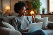 © Oleksandr - Smiling Black woman waves hello on video call. Young adult woman uses laptop on couch. Online communication, remote work, virtual meeting, telehealth session.