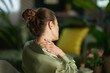 © Alliance - A woman in a green blouse reaches back to touch her neck, depicting muscle tension and the need for a mindful pause and physical relief in her home environment.