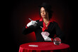 © Pixel-Shot - Young African-American magician with magic wand, cards and crystal ball on table against dark red background