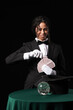 © Pixel-Shot - Young African-American magician with cards and crystal ball on table against black background