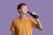 © Pixel-Shot - Tattooed young happy man with bottle of beer on purple background