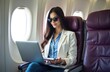 © Vadym - Woman in sunglasses works on laptop inside airplane cabin. She smiles while flying in her near window. Business traveler enjoys long flight. Comfortable journey.