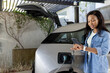 © wavebreak3 - African American woman standing beside charging EV in carport checking watch and phone, copy space