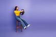 © deagreez - Young woman sits on a stool holding a steering wheel in a purple studio background