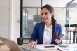 © SOMKID - Smiling Asian businesswoman in blue suit working on laptop and using smartphone in modern office.