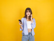 © Designerant - Concerned caucasian woman holding smartphone and touching neck with uneasy expression, reacting to confusing message or situation, wearing striped shirt, isolated yellow background with copy space.