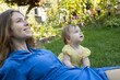 © Anna - mother and her little daughter are sitting on the grass in the garden, looking in the same direction. The closeness of generations, caring for a small child. A pleasant moment of motherhood