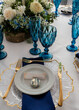 © Manuela - Elegante mesa de evento con decoración floral, copas azules y vajilla dorada en celebración sofisticada