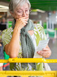 © luciano - Worried senior woman standing in a supermarket checkout area, holding and reviewing her shopping receipt beside a grocery cart, expressing concern about household expenses and rising food prices