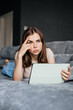 © Alena Zolot - Young woman is lying on the couch with a tablet in her hand. He looks away, frowning, thinking about something.