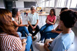 © CarlosBarquero - A diverse group of young adults and their instructor sit in a circle for an open dialogue session. They are smiling and sharing ideas in a spacious school room with a blackboard.