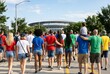 © imagezine - Rear view of diverse crowd walking along American sidewalk toward generic modern stadium on sunny summer match day