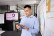 © fizkes - Project coordinator review. Confident young businessman stand near Kanban workflow model with tasks schedule on sticky notes check team work progress on digital tablet set deadlines priorities online