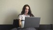 © Vlad - Young woman with thermomug sitting on cozy bed, writing email on laptop at home