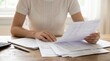 © Phaimanee - A person sitting at a wooden desk, reviewing financial documents or bills, with a calculator and pen nearby. The scene suggests someone managing personal finances or going through paperwork.