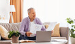 © Prostock-studio - An elderly man sits on a couch in a bright living room. He holds papers in his hands while looking at a laptop. Sunlight fills the space, creating a warm atmosphere for work.