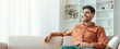 © Prostock-studio - A young man sits comfortably on a couch in a bright room. He has a smile on his face and seems to enjoy his leisure time. Sunlight is streaming through the window, creating a warm atmosphere.