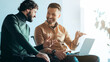 © Prostock-studio - Two men sit next to each other in an office setting. They are engaged in conversation while looking at a laptop. Sunlight filters through a window, illuminating the space.