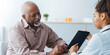 © Prostock-studio - An elderly man sits in a medical office talking to a healthcare worker. They are looking at a tablet together. The healthcare worker points to something on the screen while explaining.