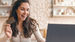 © Prostock-studio - A woman is sitting at a desk with a laptop in front of her. She holds a credit card in her right hand and has a happy expression. She appears engaged in online shopping from her home.