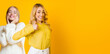 © Prostock-studio - Two women stand in front of a yellow backdrop. One woman smiles and poses with a thumbs up while the other appears happy in the background. The setting is bright and cheerful.
