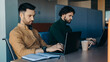 © Prostock-studio - Two men are focused on their laptops while sitting at a wooden desk in an office. They are working quietly with a notebook and a cup of coffee nearby.