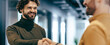 © Prostock-studio - Two men are shaking hands in an office setting. They are smiling and appear to be pleased with their discussion. The background shows a modern workspace with bright lighting.