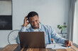 © Daniel - A young man sits at a wooden table in a home office, staring at his laptop with a puzzled expression while sorting paperwork and taking notes, conveying frustration and concentration during work