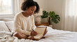 © Mukhliez - Black woman writing in journal on messy bed in morning sunlight